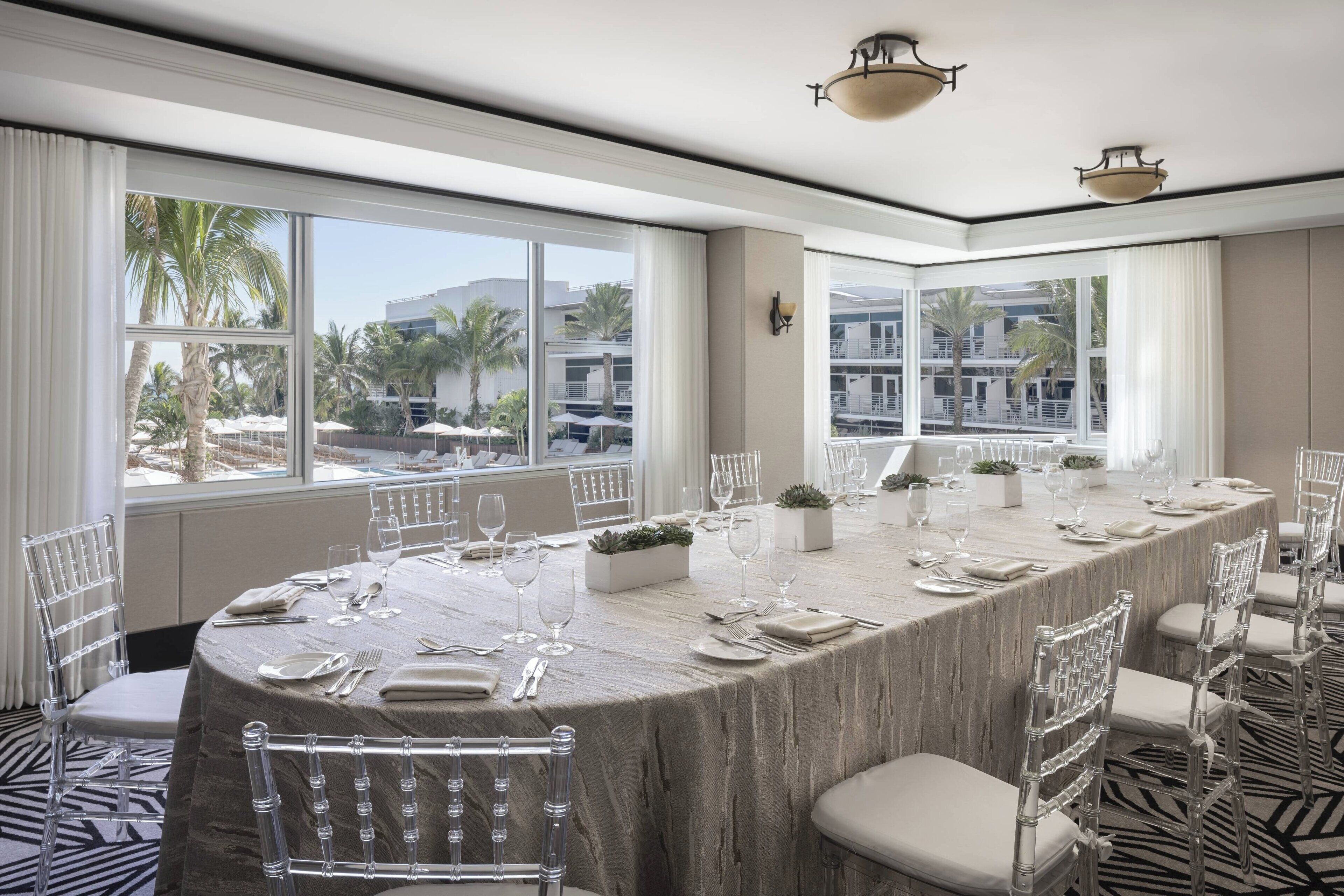 Une grande salle de conférence dotée d’une table en marbre préparée pour une réunion, de grandes fenêtres offrant une vue sur des palmiers et des immeubles, et de chaises modernes à structure métallique.
