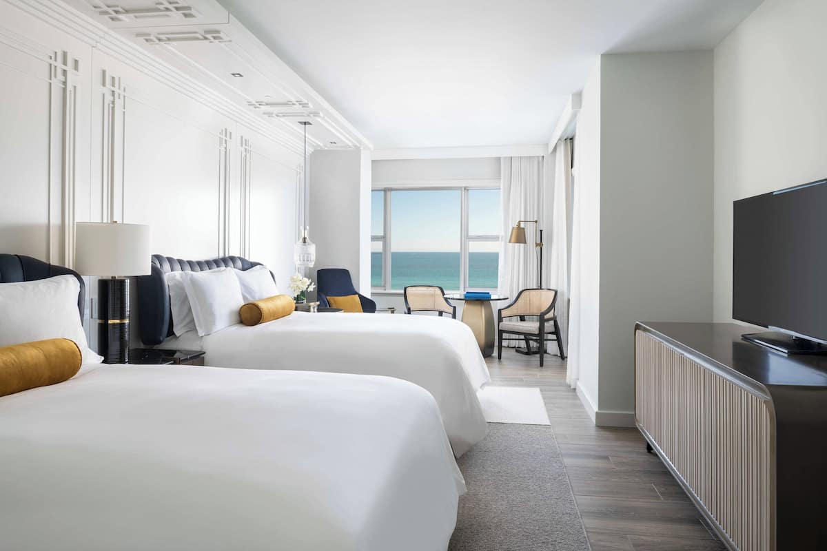 A hotel room with two beds, a desk, a chair, a television, and a view of the sea.