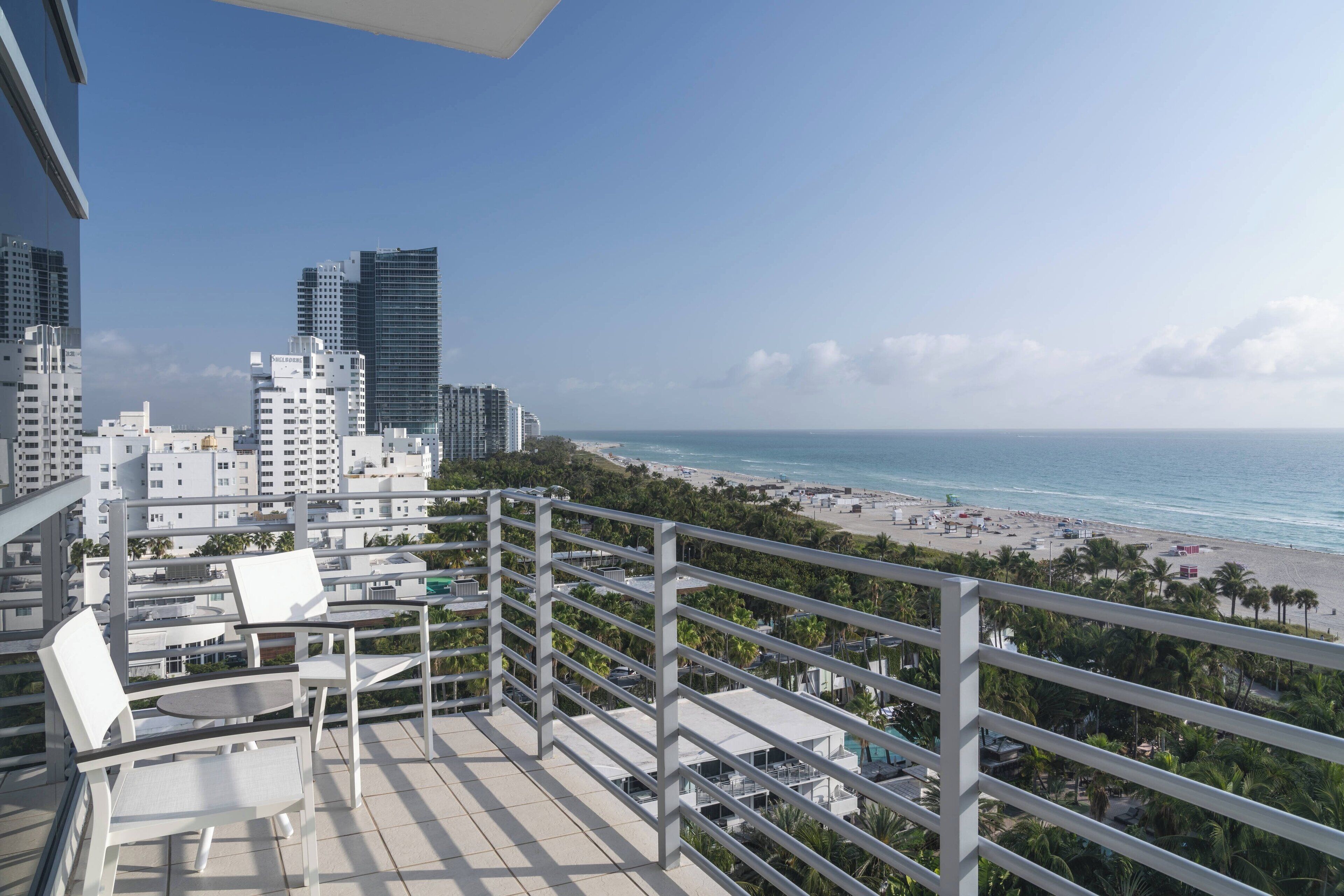 Ein Balkon mit weißen Stühlen und einem Tisch, der Blick auf einen Strand mit Palmen und eine Skyline im Hintergrund bietet.