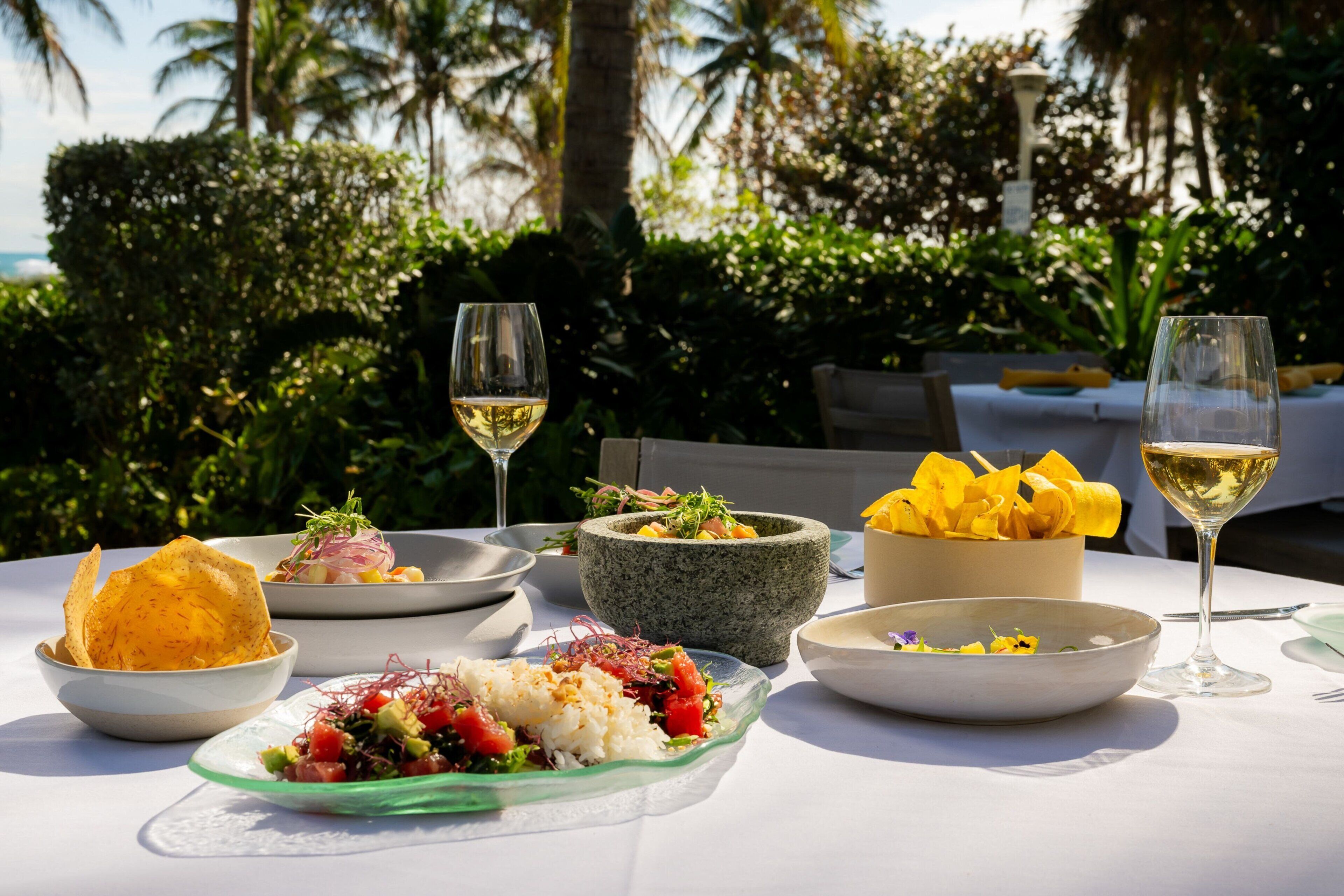A table set with various dishes and drinks, surrounded by lush greenery and palm trees.
