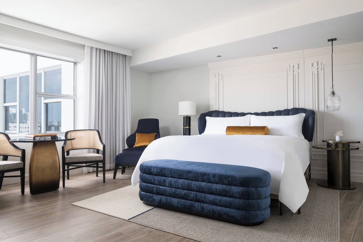A modern hotel room with a large bed, a seating area with chairs, a small table, and a view of the city through large windows.