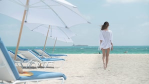 Uma praia com fileiras de espreguiçadeiras e guarda-sóis, céu claro e o oceano ao fundo.