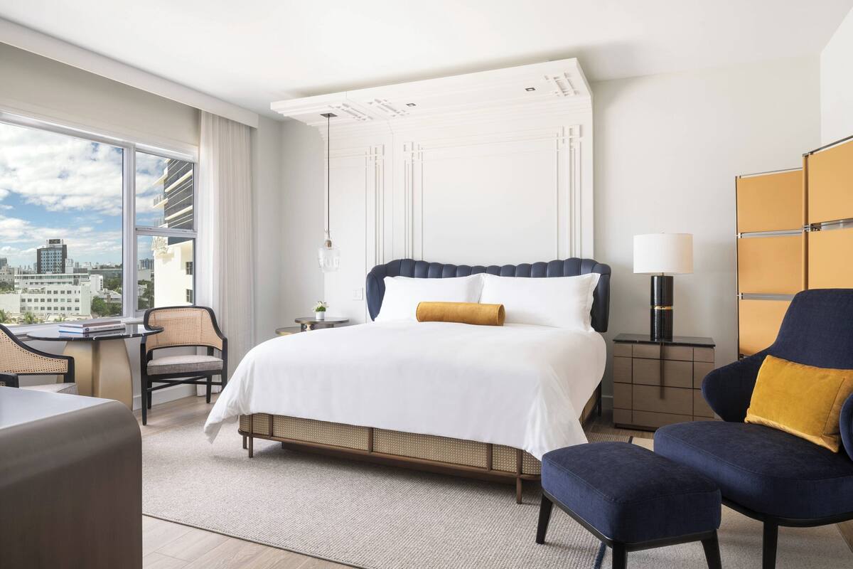 A modern hotel room with a large bed, a desk, a chair, and a view of the city.