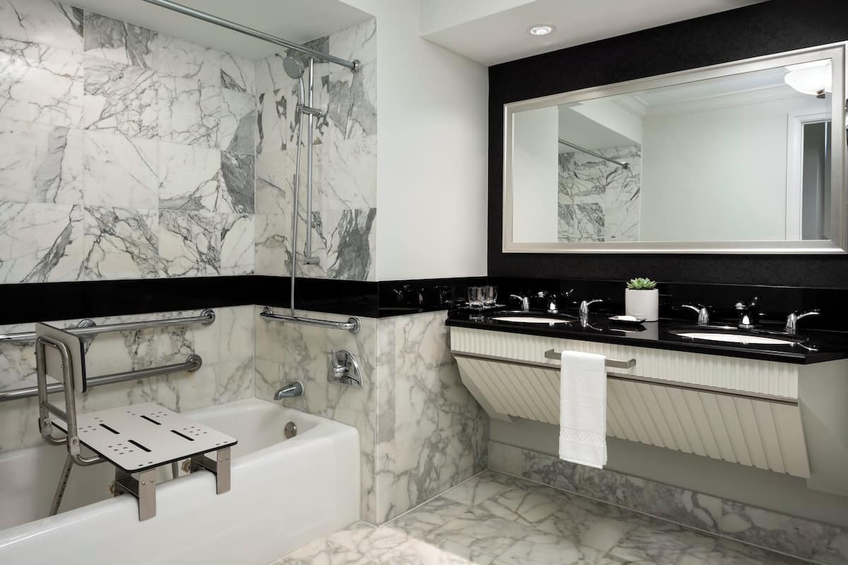 A modern bathroom with a marble shower, a bathtub with a lift, a large mirror, and a black countertop with a sink and a potted plant.