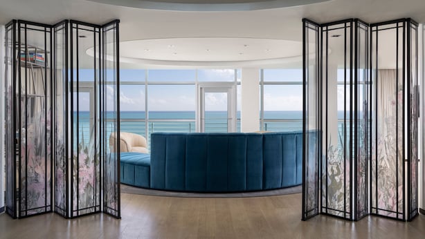 Quarto moderno com sofá azul curvo, uma janela grande com vista para o mar e teto com iluminação embutida.