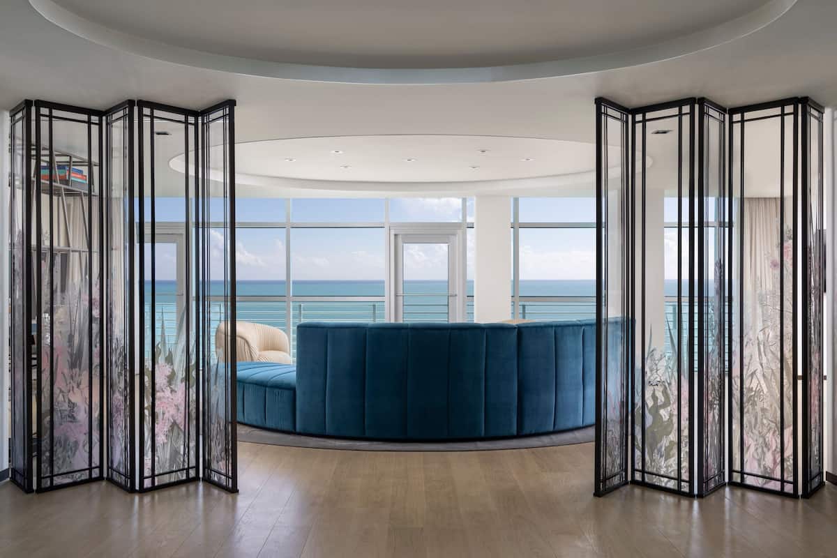 A modern room with a curved blue sofa, a large window offering a view of the sea, and a ceiling with recessed lighting.