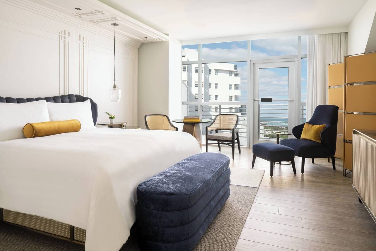 A hotel room with a large bed, a small table with chairs, a view of the city through large windows, and a balcony.