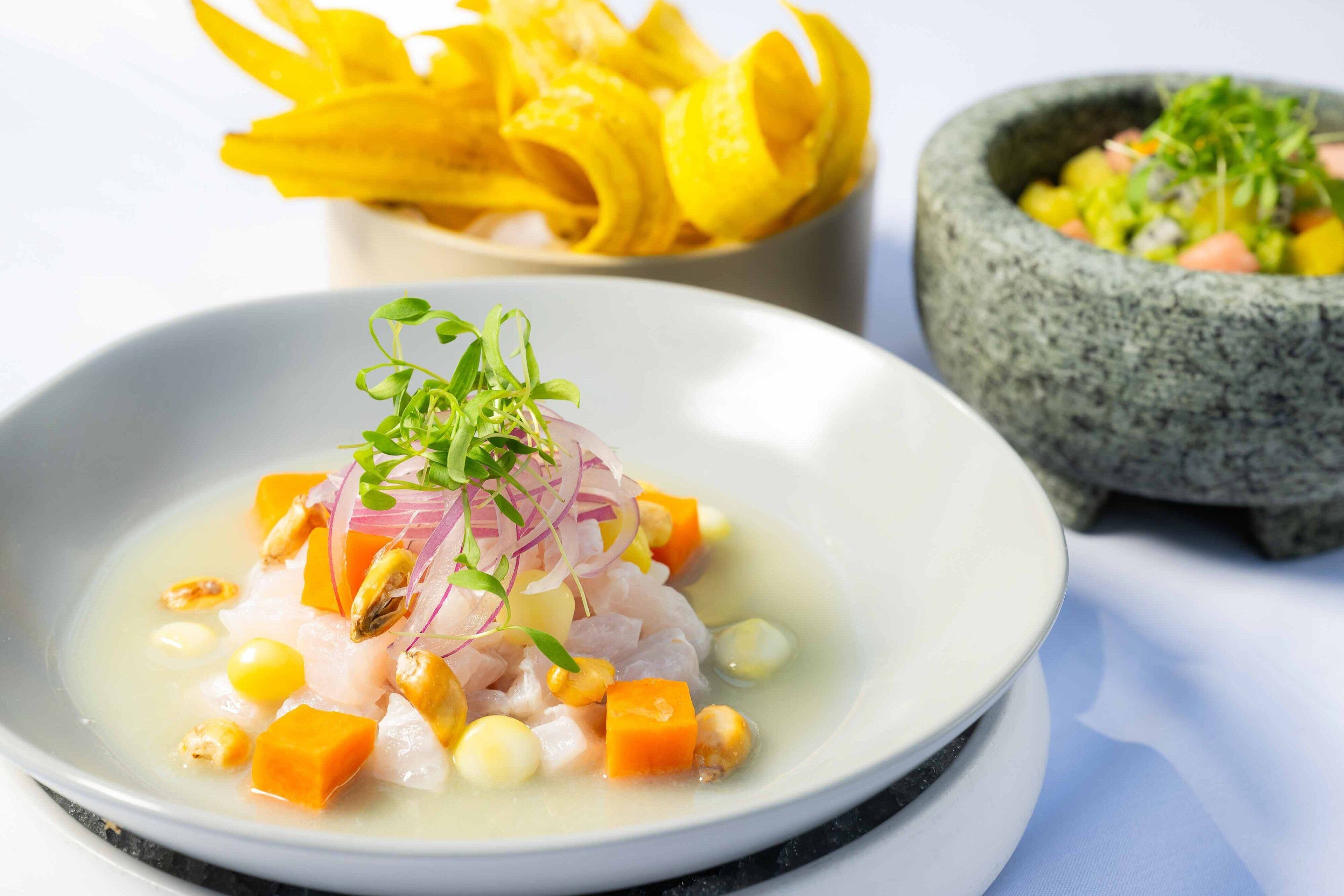 A dish with various ingredients, a bowl of yellow fruit, and a stone mortar with a green herb garnish.