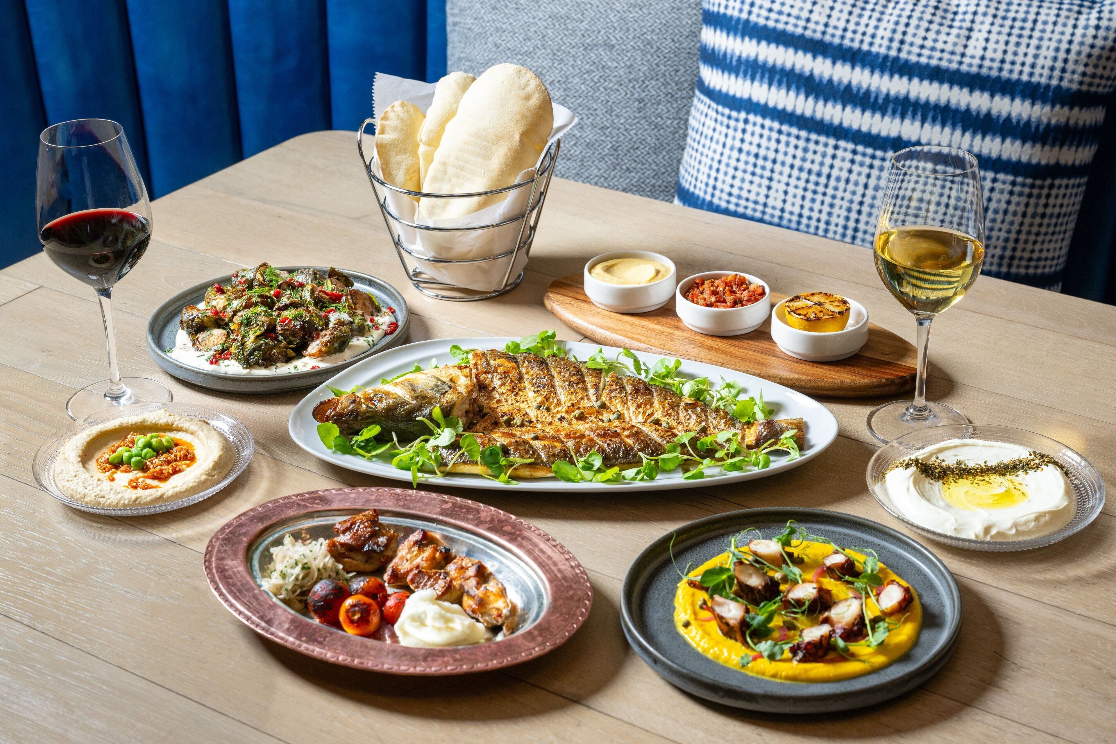 A dining table with various dishes, including a whole grilled fish, bowls of hummus, and a glass of red wine.