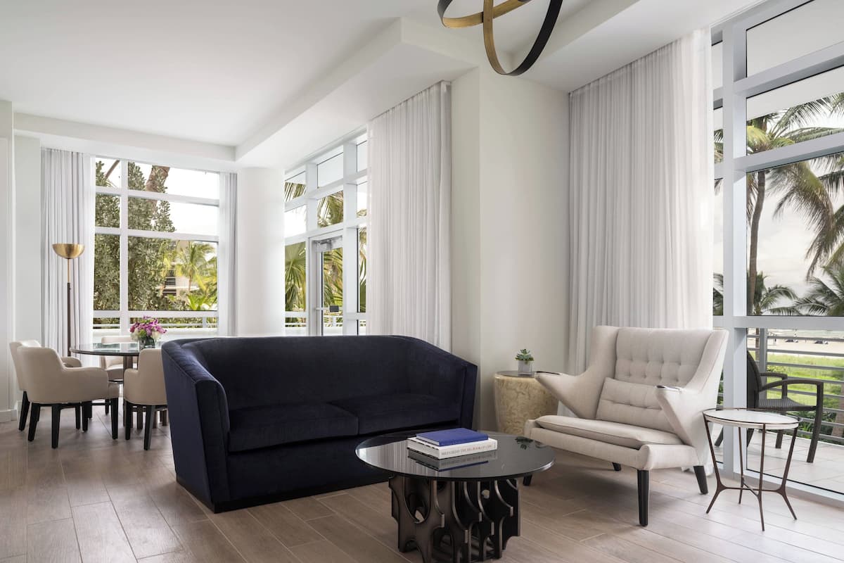 A spacious living room with a dark blue sofa, a round coffee table, and a light-colored armchair. There is a dining area with a table and chairs, and large windows with curtains.