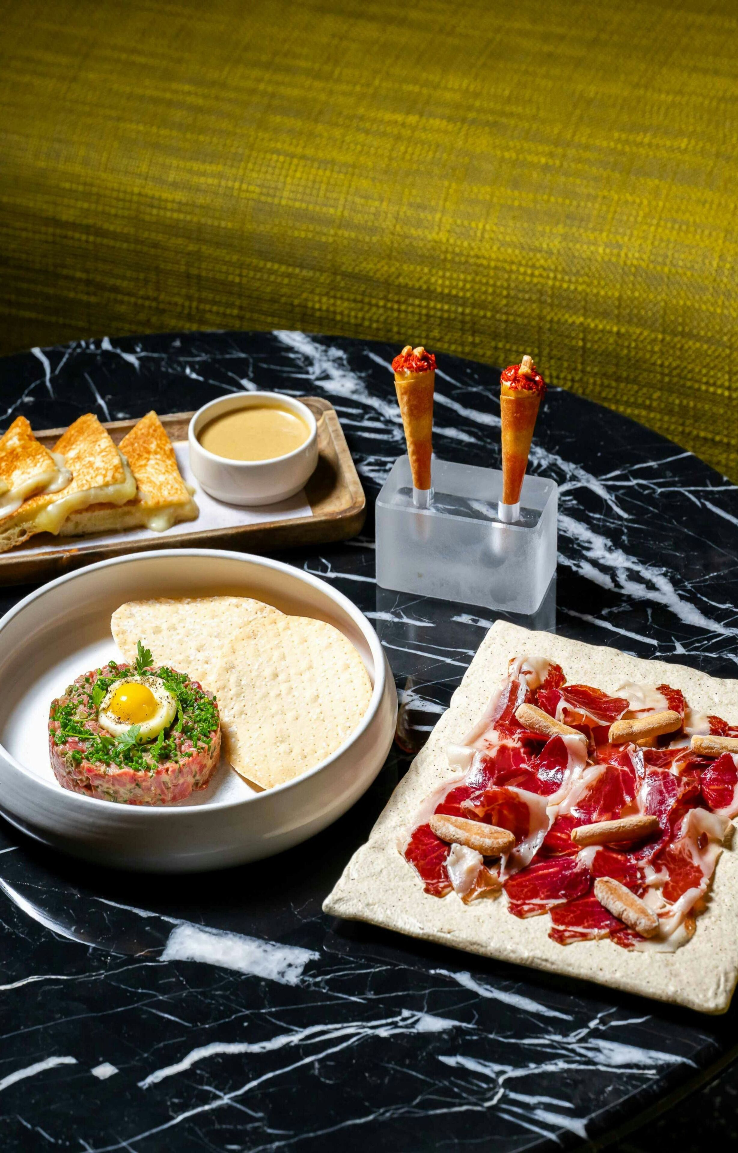 A marble table with a variety of dishes including a steak tartare, a pizza with red meat, and a sandwich.