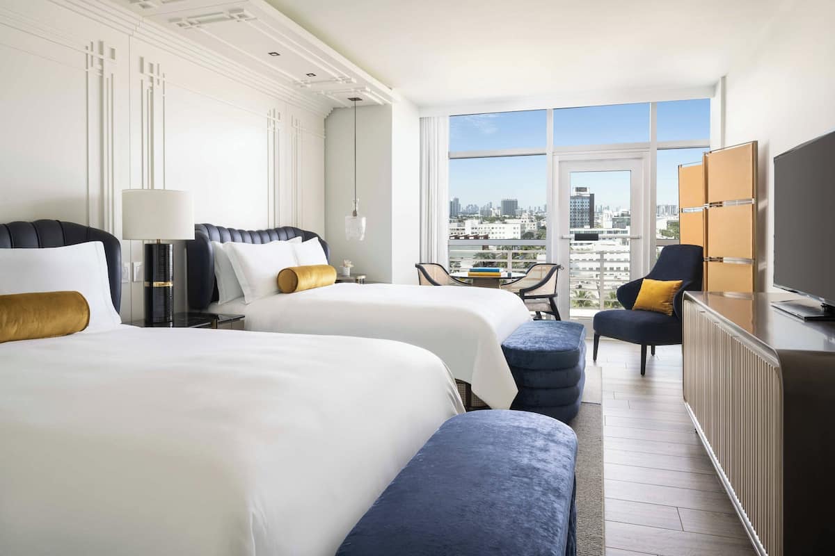 A hotel room with two beds, a TV, a desk, and a view of the city.