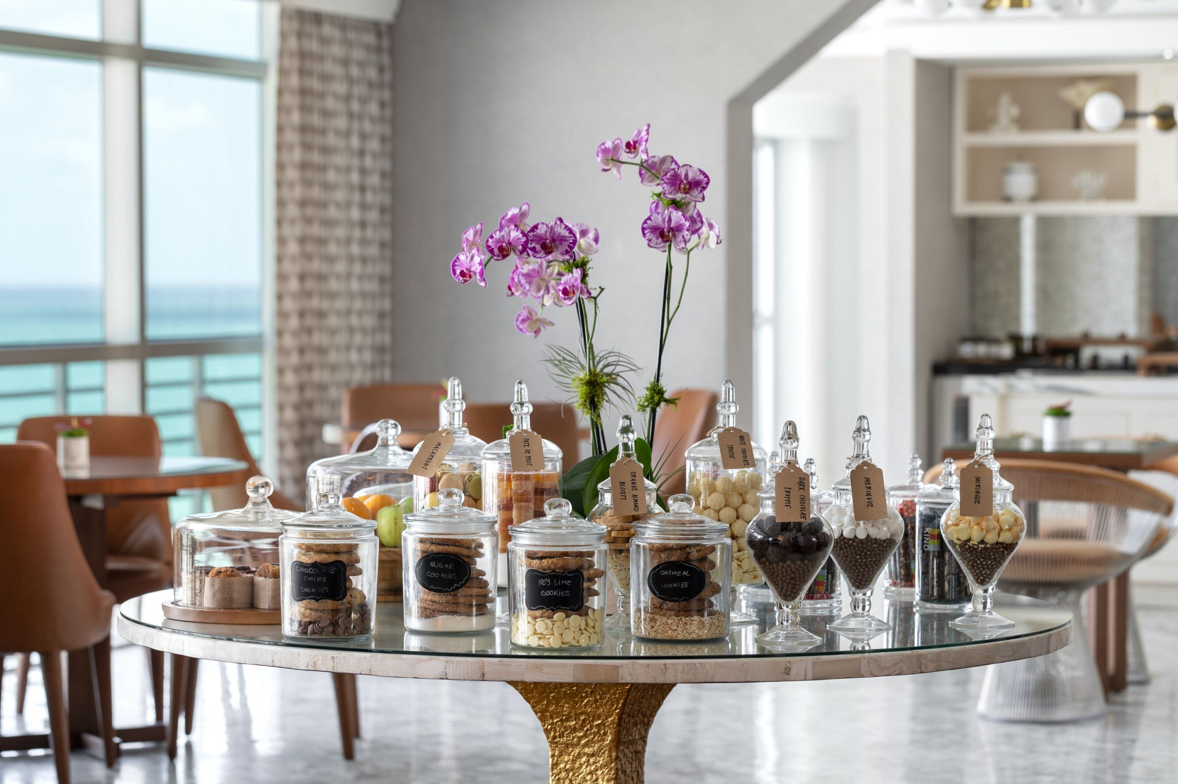 Une table à manger ornée de bocaux en verre contenant divers aliments, un vase avec des fleurs violettes, et une vue sur l’océan par de grandes fenêtres.