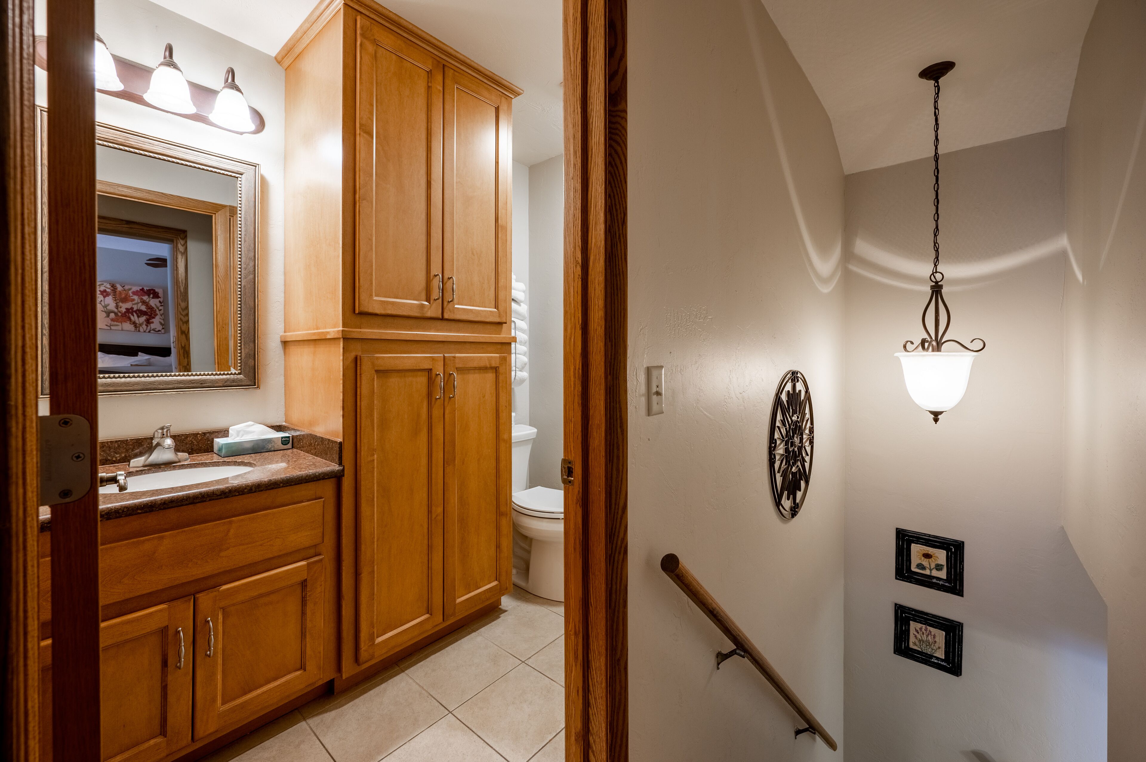 A bathroom with wooden cabinets, a sink, and a toilet. There is a mirror with lights above it, and a wall-mounted light fixture.