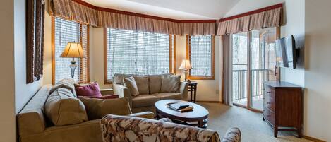 Ein Wohnzimmer mit Sofa, Sesseln und Couchtisch, einem an der Wand befestigten Fernseher sowie einem Fenster mit Vorhängen.