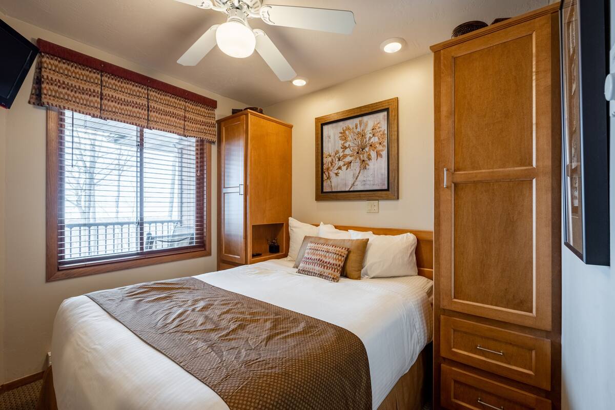Quarto com cama, ventoinha no teto, janela com persianas, guarda-roupa de madeira e um quadro na parede.