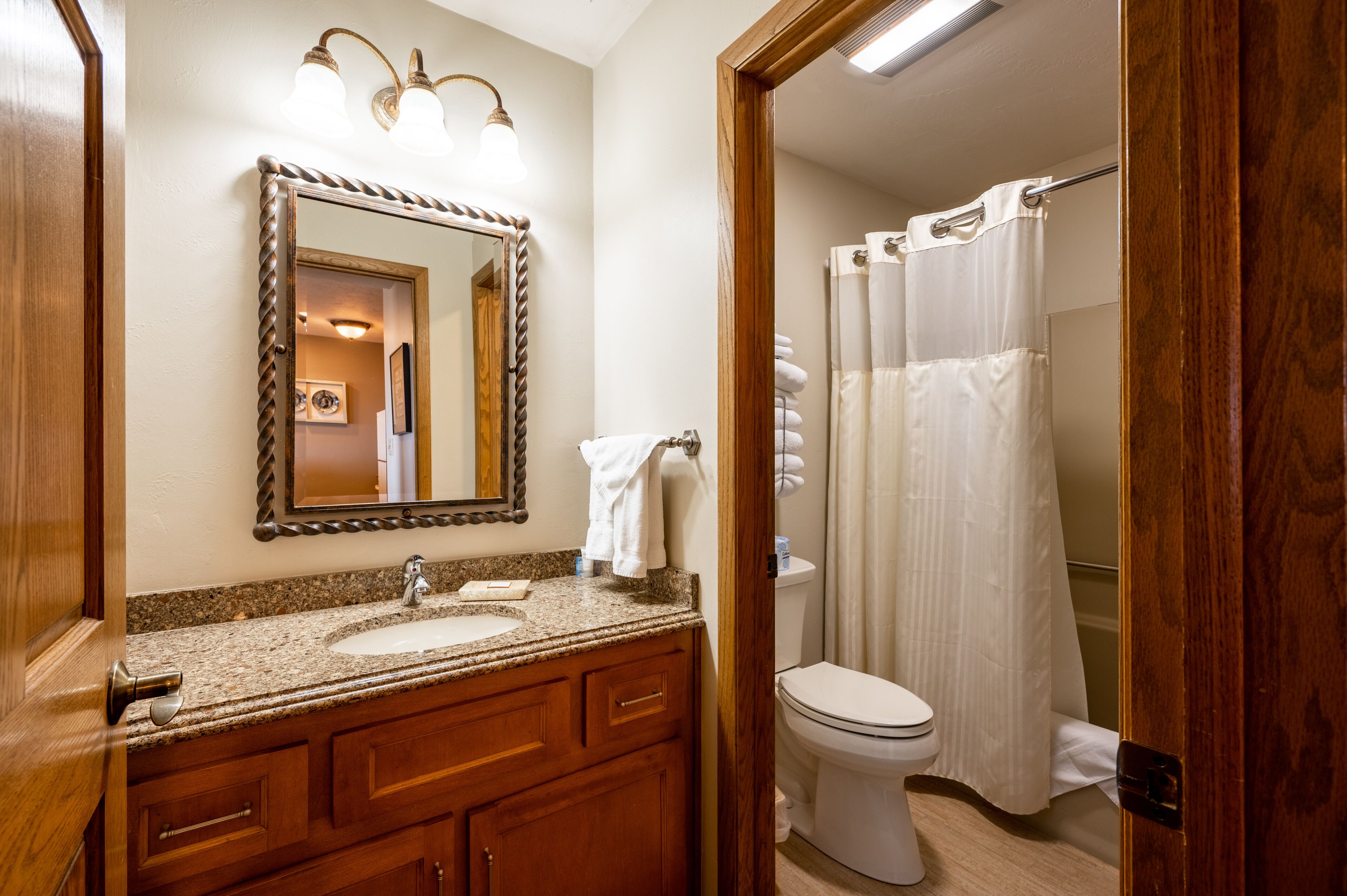 A bathroom with a granite countertop sink, wooden vanity, a mirror with a twisted frame, a toilet, and a shower with a white curtain.
