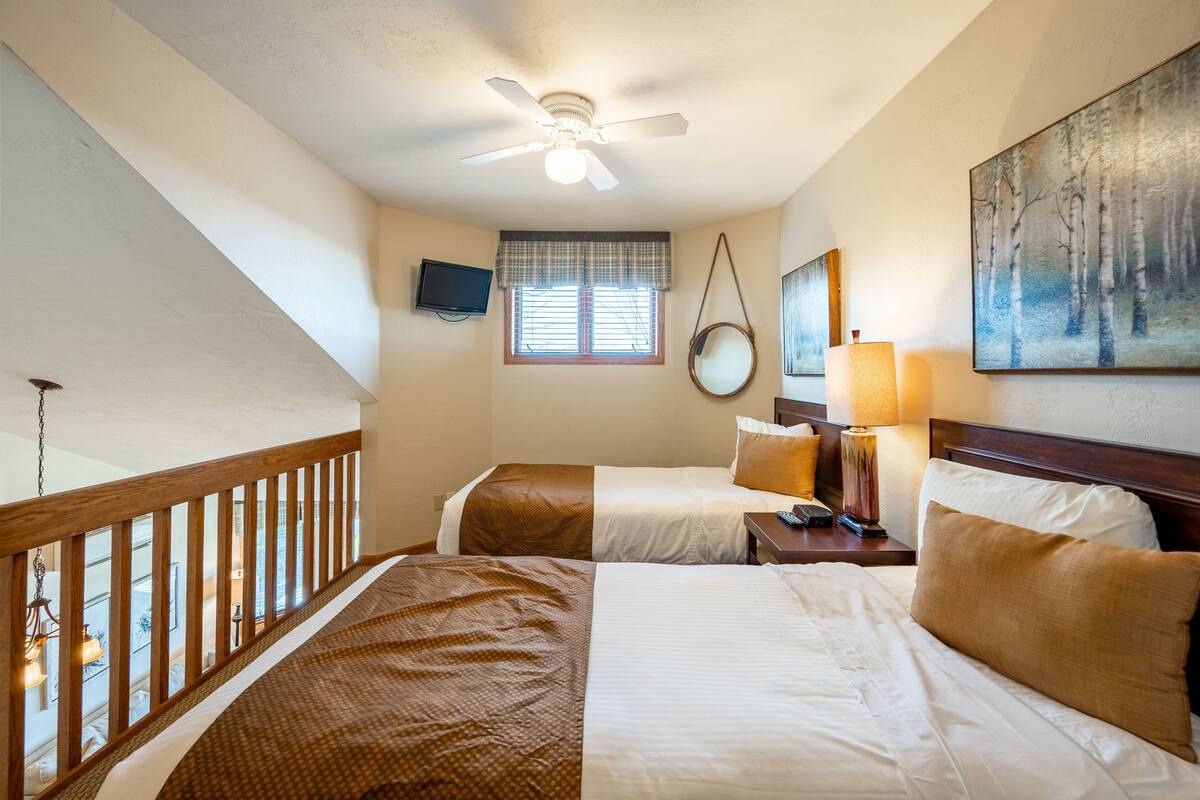 Quarto de hotel com duas camas, um ventilador de teto, televisão, janela com persianas, um quadro e um abajur.