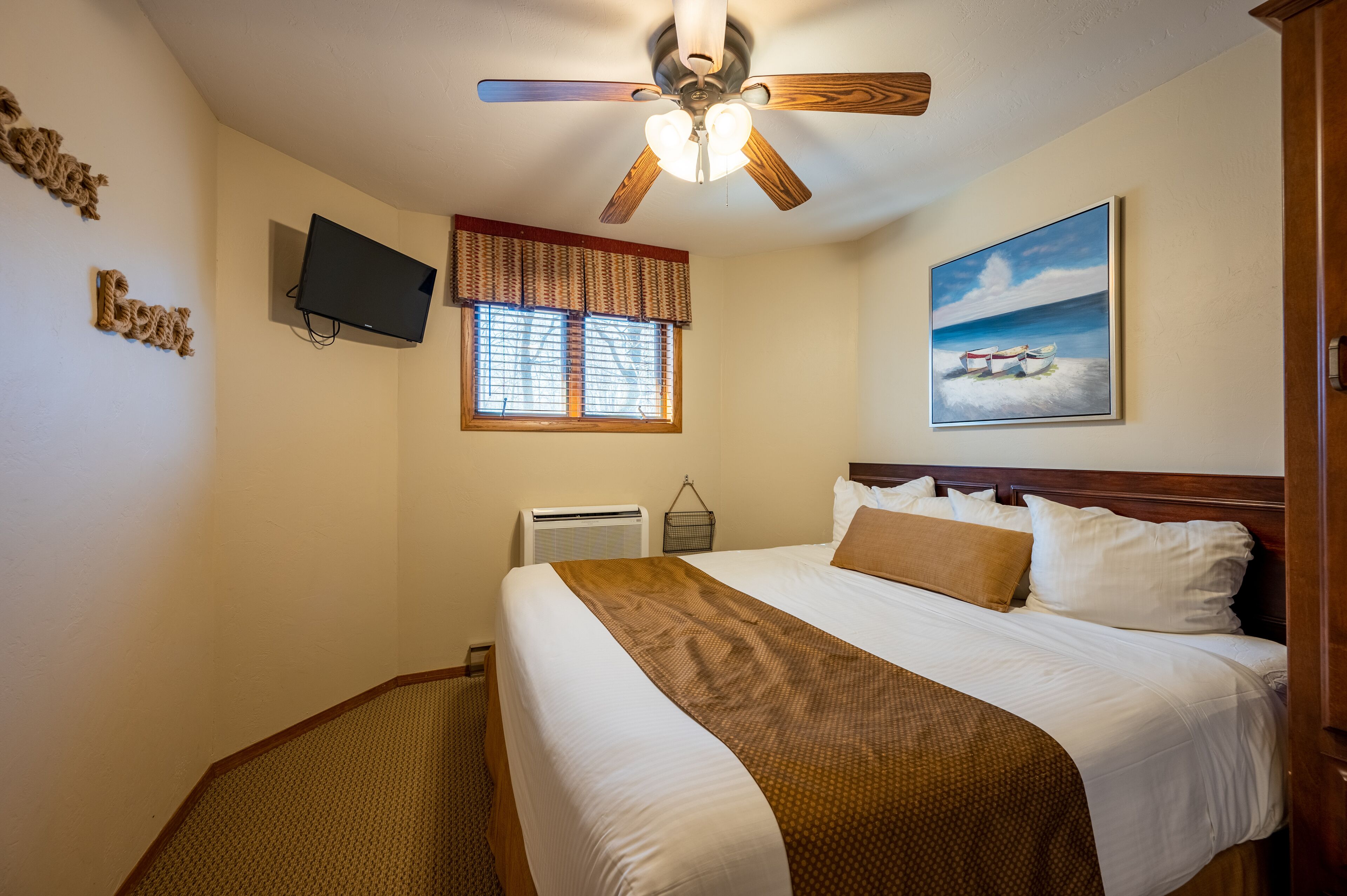 A hotel room with a bed, a ceiling fan, a TV, a window with blinds, a painting, and a wall-mounted air conditioner.