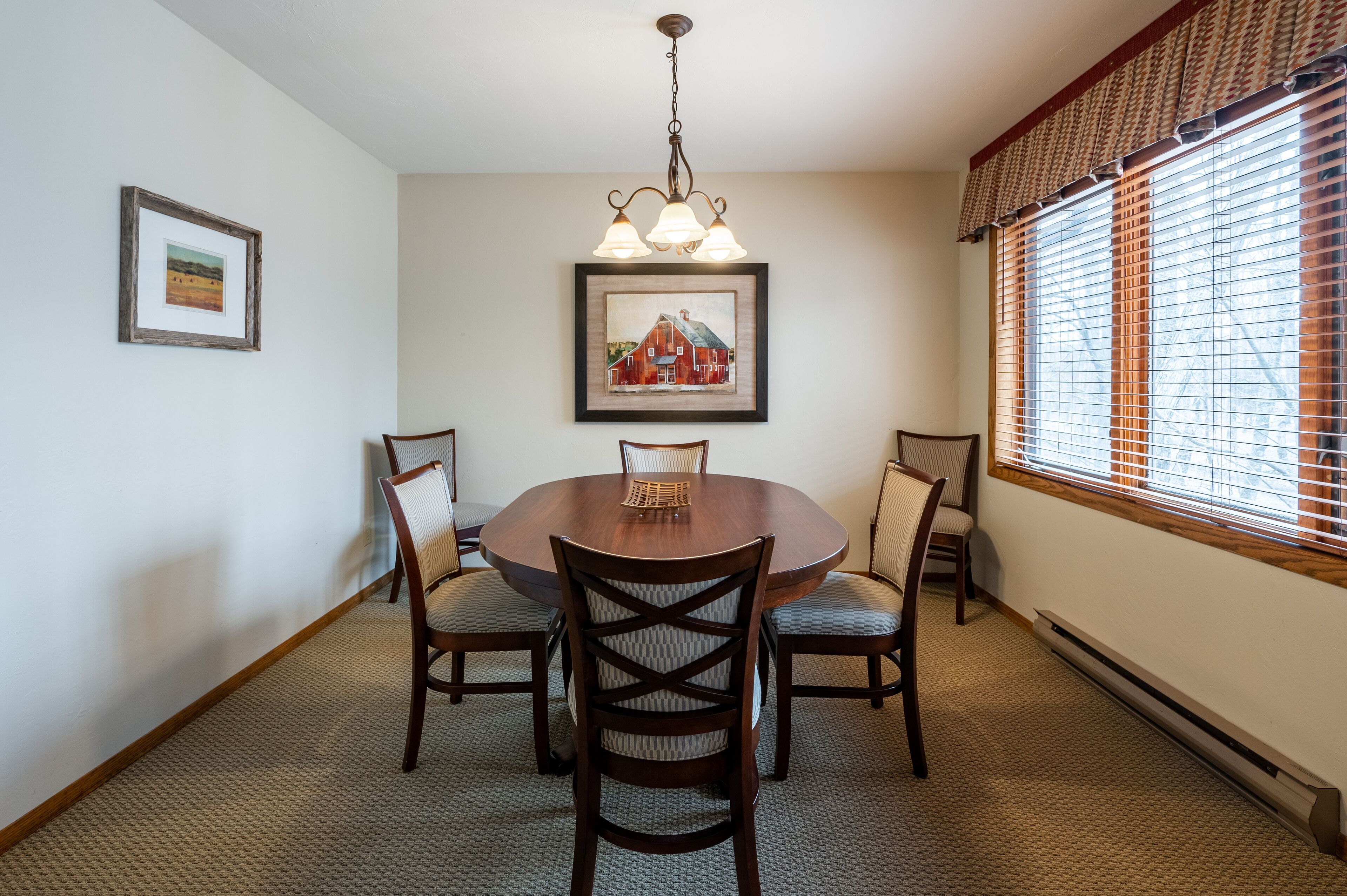 A dining room with a round table, four chairs, a chandelier, and two framed pictures on the walls.