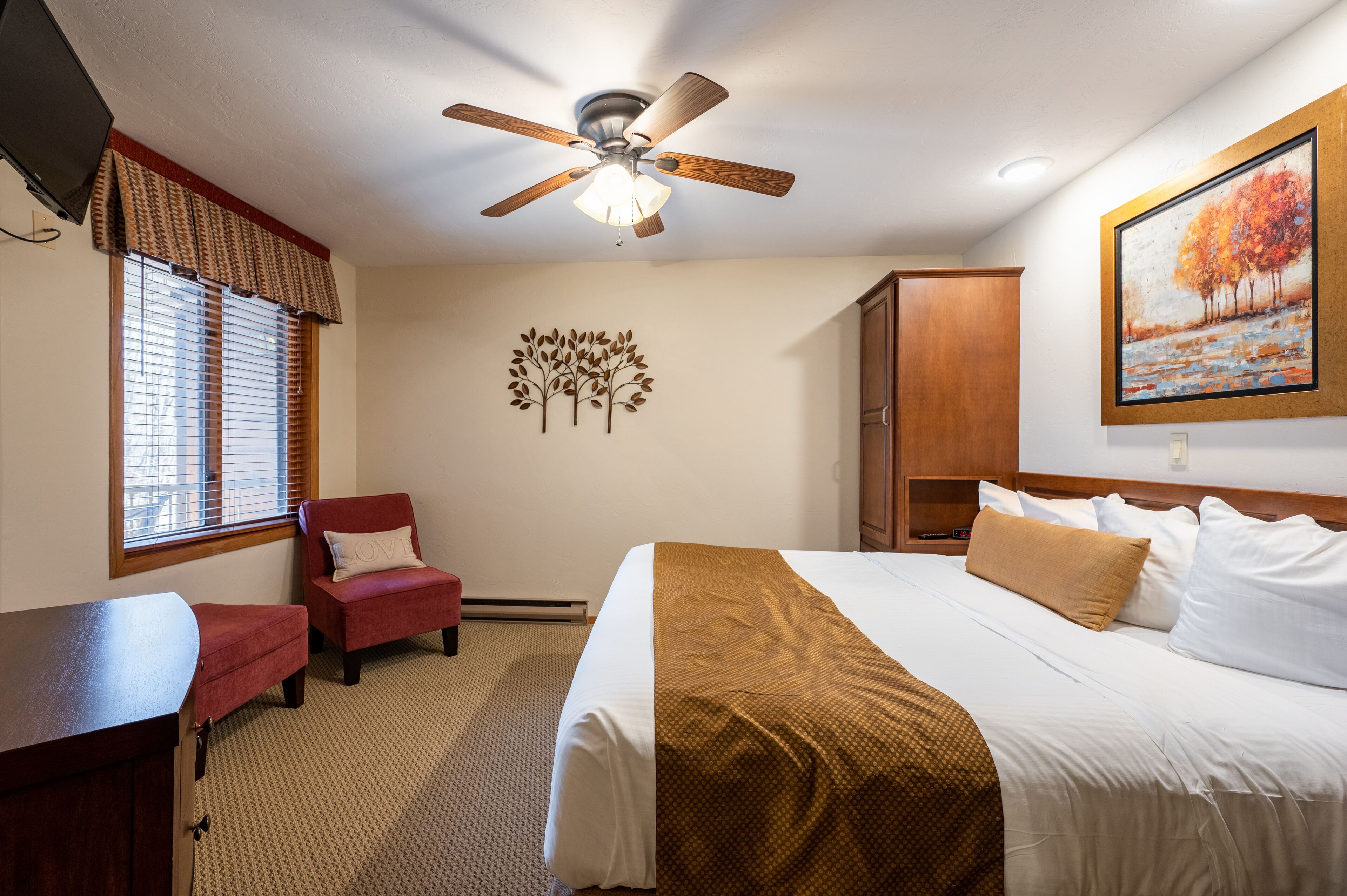 A hotel room with a bed, a desk, a chair, a ceiling fan, a painting, and a window with blinds.