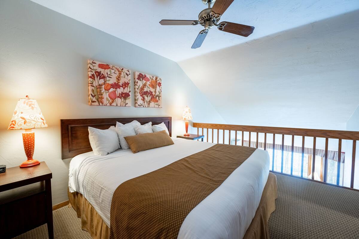 Quarto de hotel com cama, duas luminárias de cabeceira, ventilador de teto e varanda com guarda-corpo.