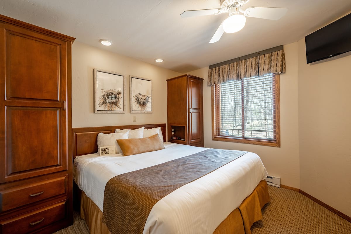 Quarto de hotel com cama grande, guarda-roupa de madeira, ventoinha no teto, obras de arte na parede e janela com persianas.