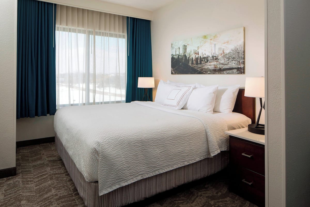 A hotel room with a large bed, two bedside tables, a nightstand, a window with curtains, and a cityscape painting on the wall.
