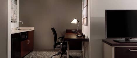 A hotel room with a kitchenette, a desk with a lamp, a chair, and a TV.