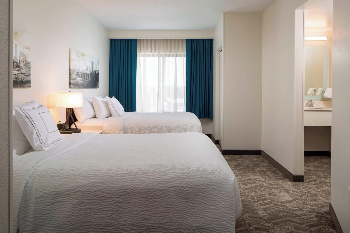 A hotel room with two beds, a window with blue curtains, a lamp, and a bathroom visible through an open door.