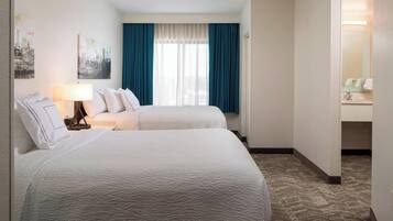 A hotel room with two beds, a window with blue curtains, a lamp, and a bathroom visible through an open door.