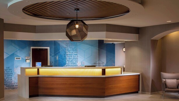 A reception area with a wooden reception desk, a geometric wall design, and a modern ceiling light fixture. - Springhill Suites By Marriott Chicago Lincolnshire (Lincolnshire)