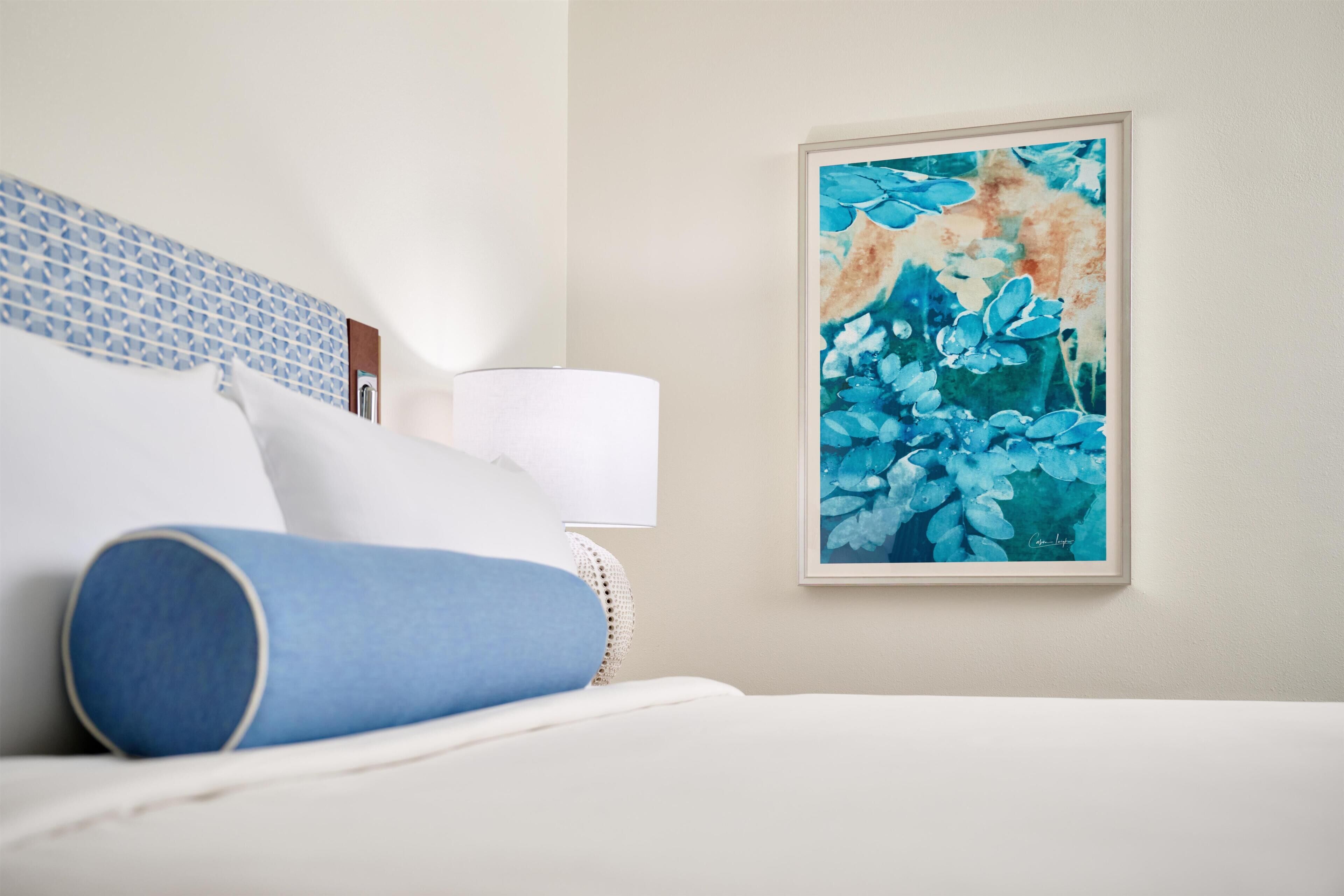 A neatly made bed with a blue pillow, a white pillow, and a white blanket. There is a framed painting of blue flowers on the wall.