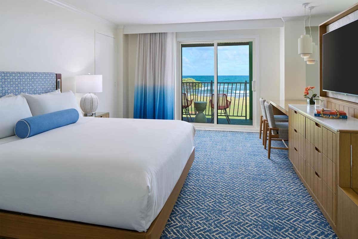 A hotel room with a bed, a desk, a TV, a balcony with a view of the ocean, and a patterned carpet.