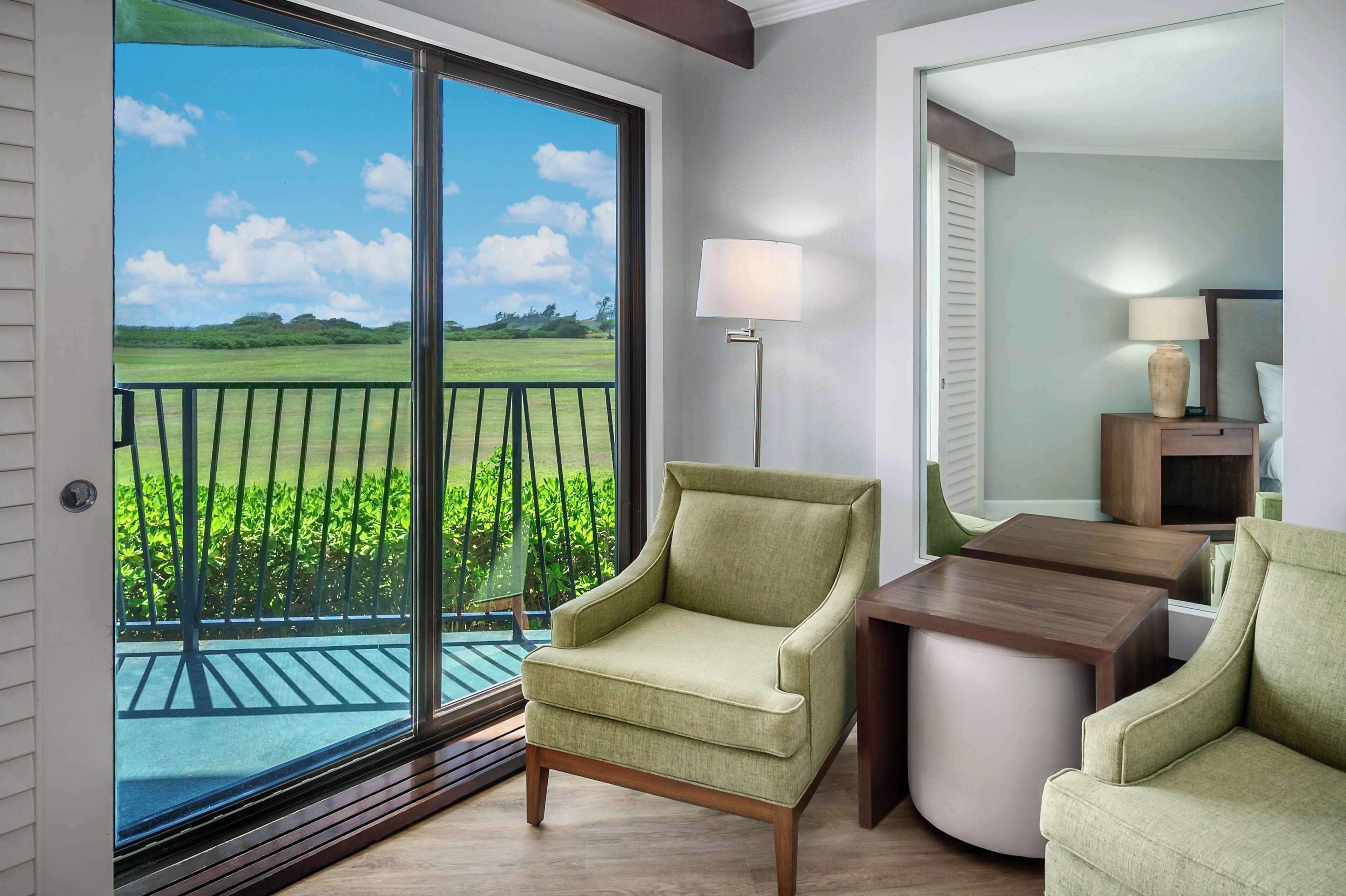 A room with a view of a green landscape through a large window, a green armchair, a small round table, and a lamp.