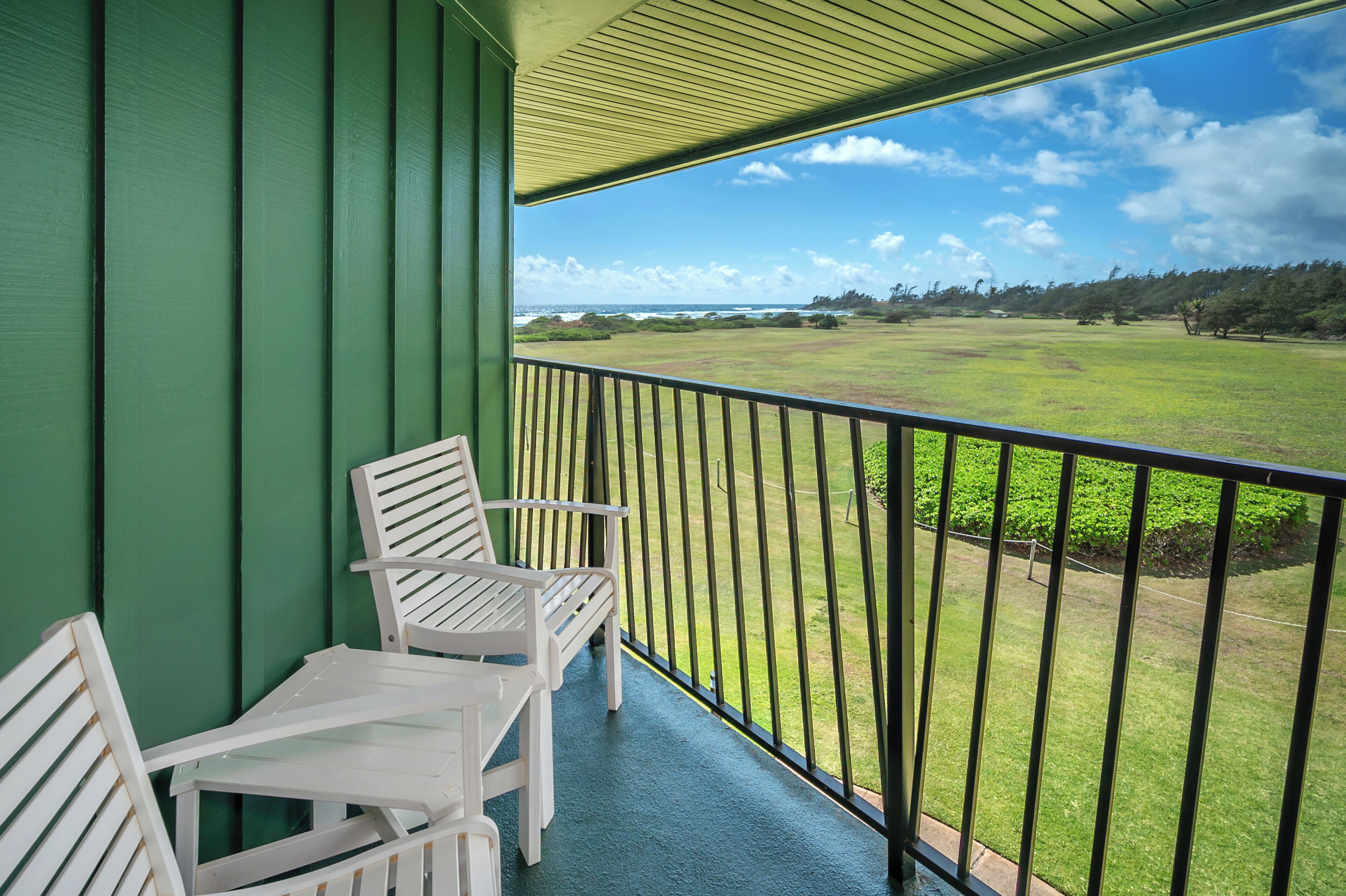 Ein Balkon mit zwei weißen Stühlen und einem Tisch, der auf ein grünes Feld und eine ferne Landschaft blickt.