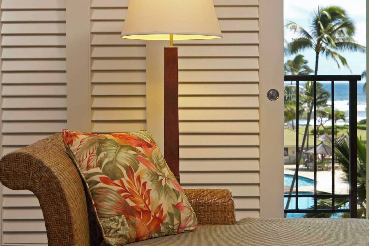 A room with a bed, a lamp, and a view of palm trees and a pool.