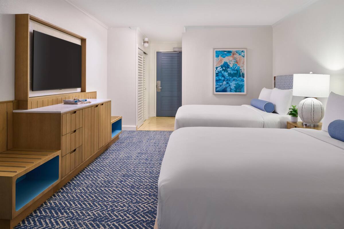 A hotel room with a TV, two beds, a painting, a lamp, and a small plant.