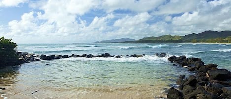 A beach with clear water, rocky shoreline, and distant mountains under a partly cloudy sky.