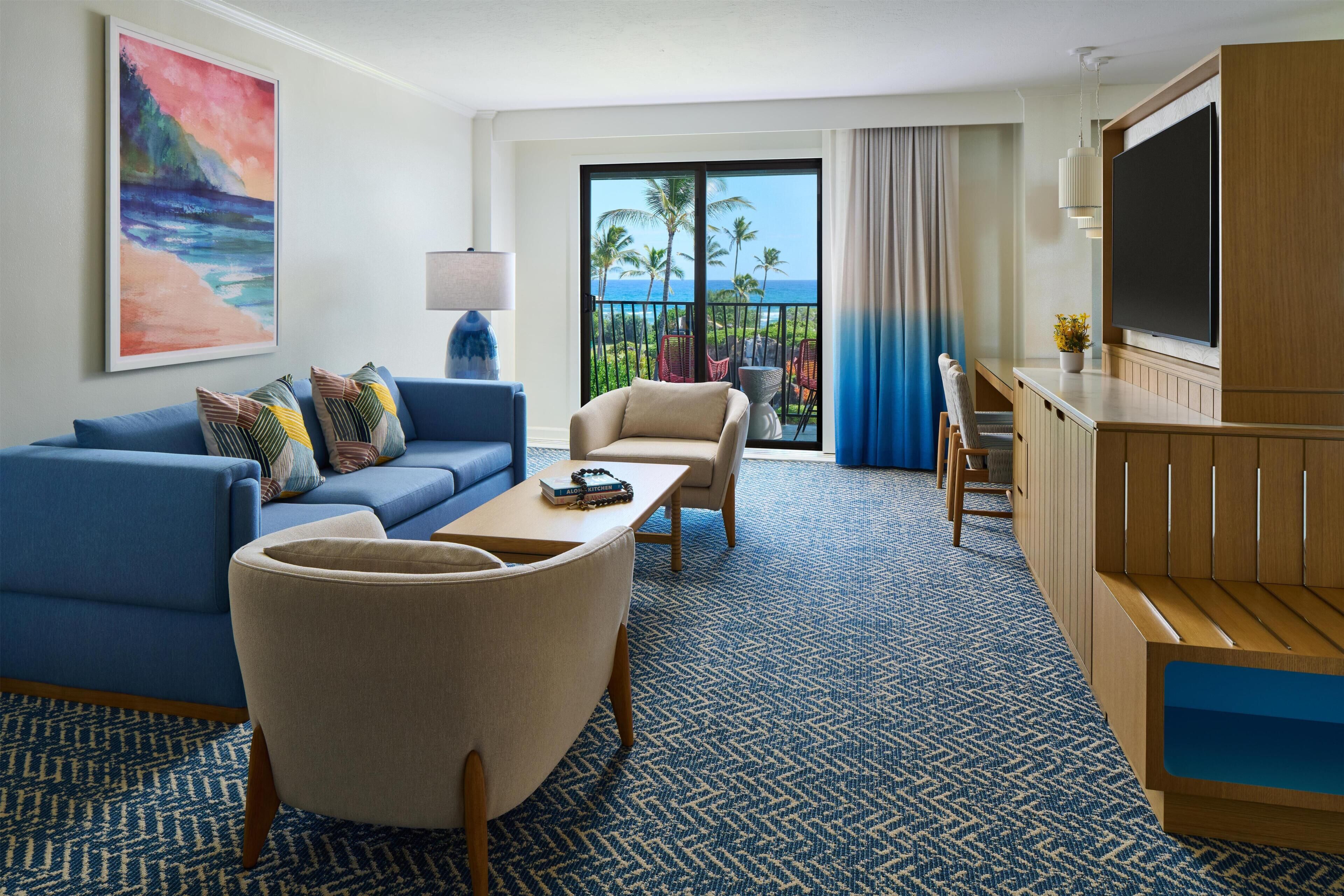 A hotel room with a blue sofa, a beige armchair, a coffee table, a TV, and a balcony with a view of palm trees and the ocean.