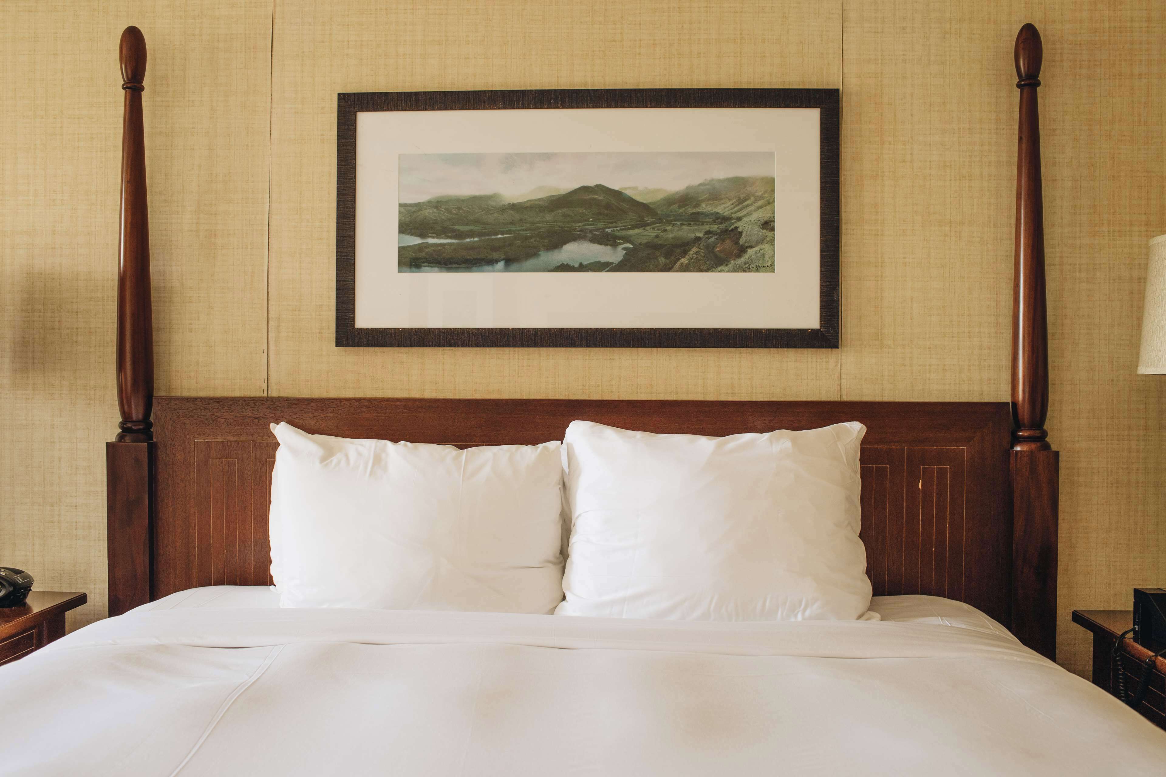 A neatly made bed with white linens and two pillows, a wooden headboard, and a framed landscape painting above it.