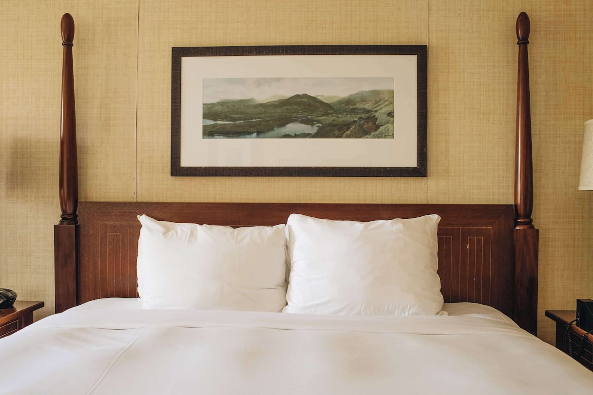 A neatly made bed with white linens and two pillows, a wooden headboard, and a framed landscape painting above it.