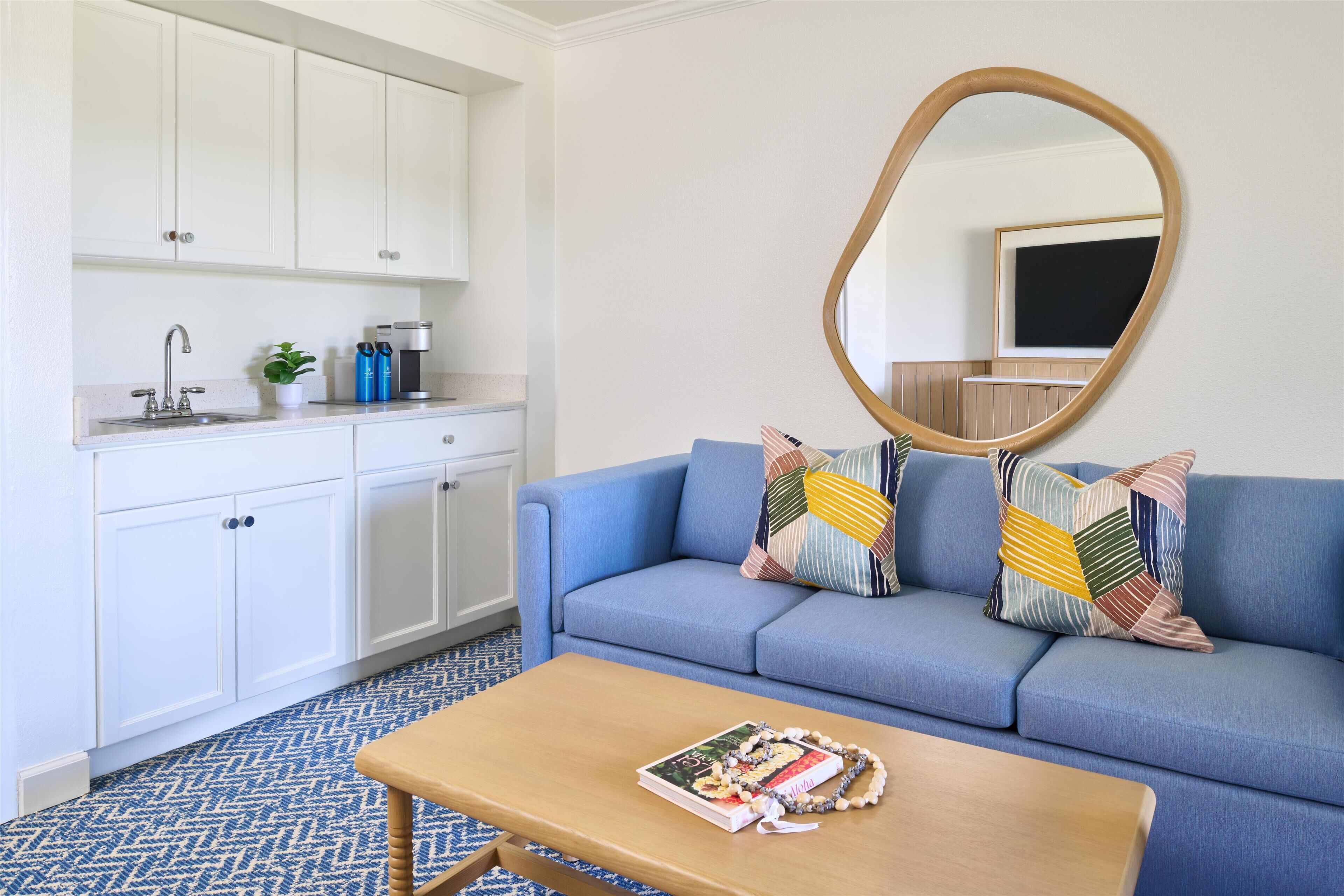 Uma sala de estar moderna com um sofá azul, uma mesa de centro de madeira e uma área de cozinha com armários brancos e uma pia.