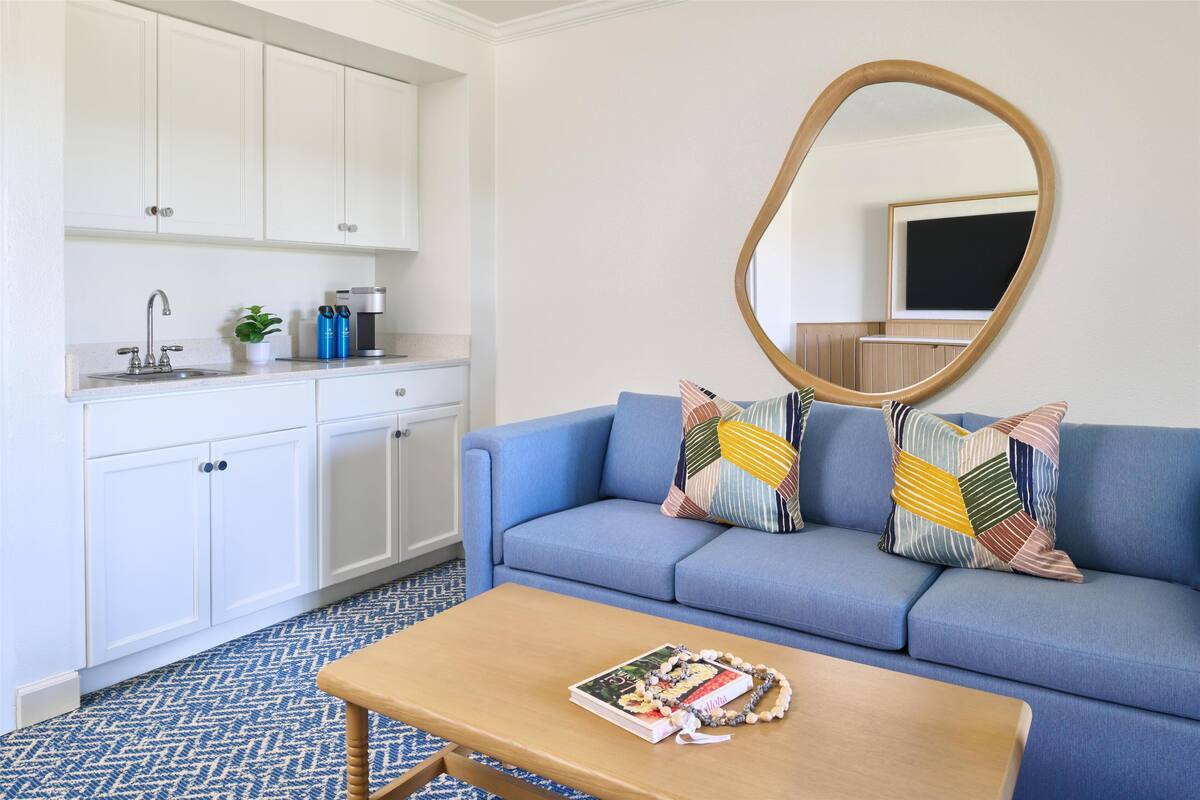 A modern living room with a blue sofa, a wooden coffee table, and a kitchen area with white cabinets and a sink.