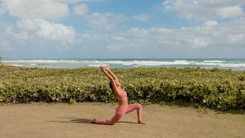 A person performing a yoga pose on a grassy area with a view of the ocean and a hedge in the background.
