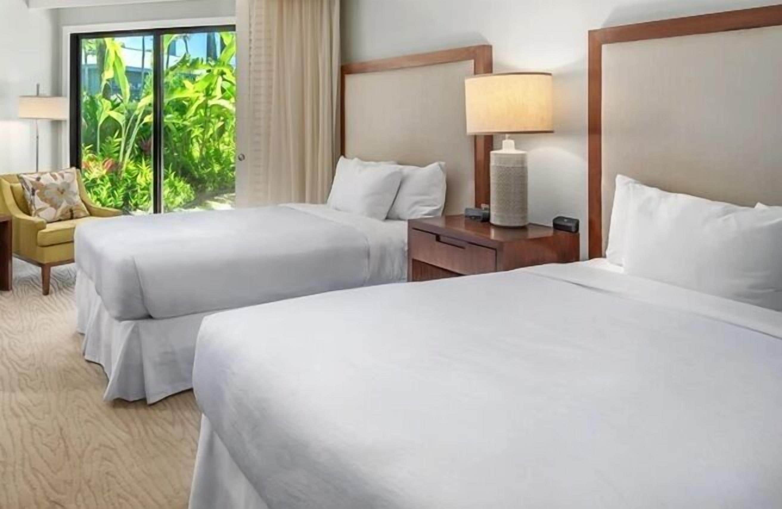 A hotel room with two beds, a nightstand with a lamp, a chair, and a view of greenery outside.