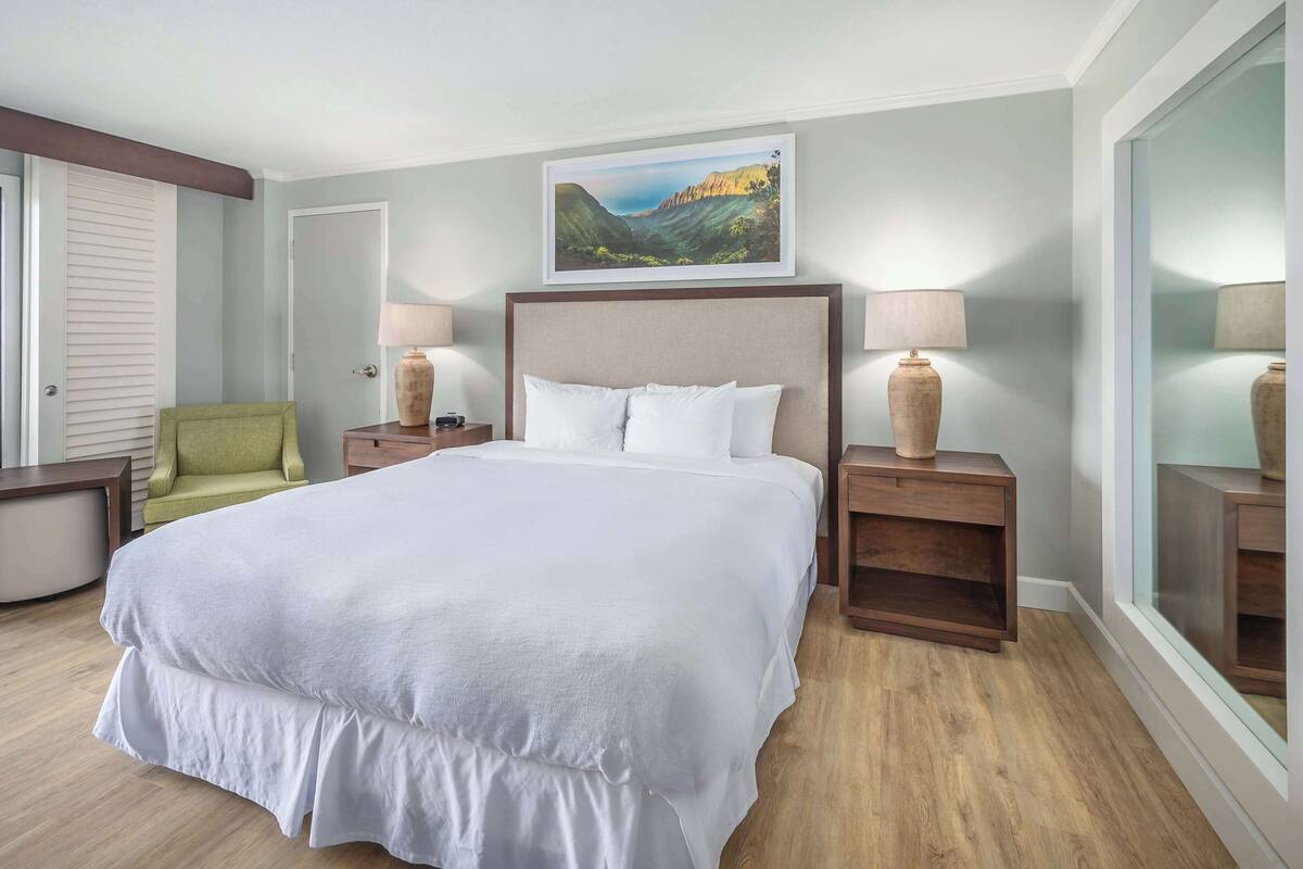 A hotel room with a large bed, two bedside tables, a green armchair, and a painting on the wall.