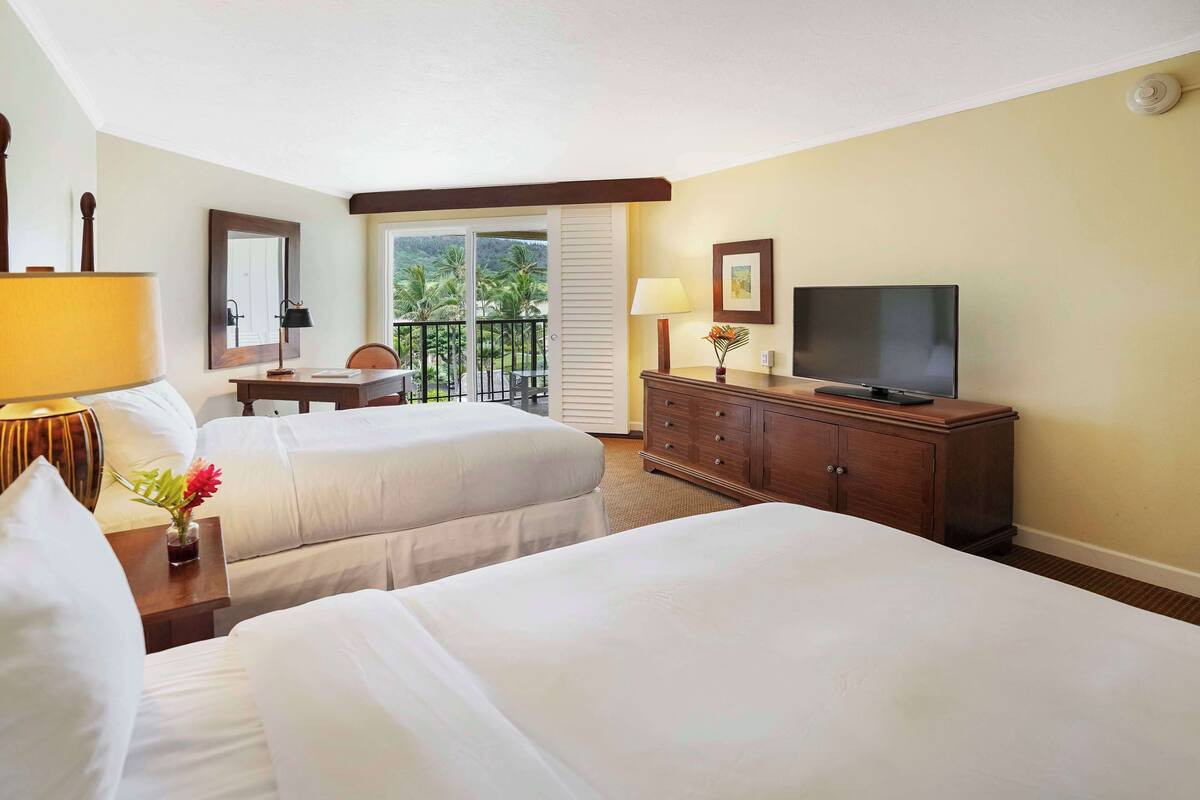 A hotel room with two beds, a desk, a television, and a balcony with a view of palm trees.