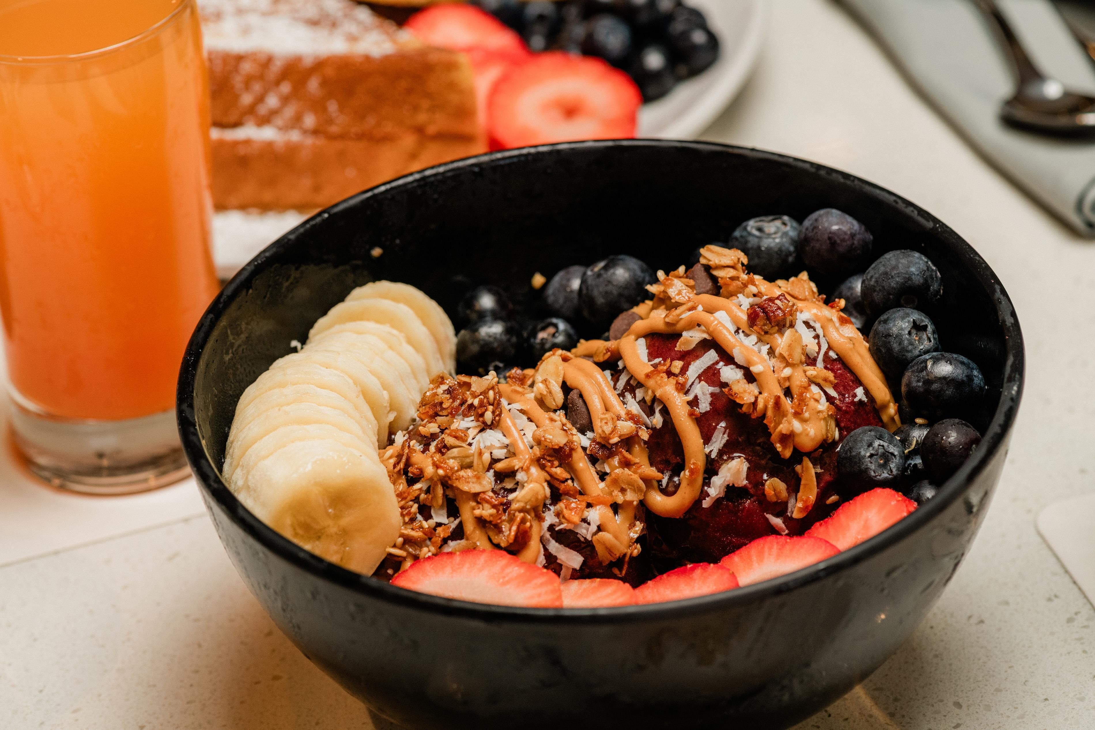 Un tazón de ensalada de frutas con plátanos, fresas y arándanos, un vaso de jugo de naranja y una rebanada de pan al lado.