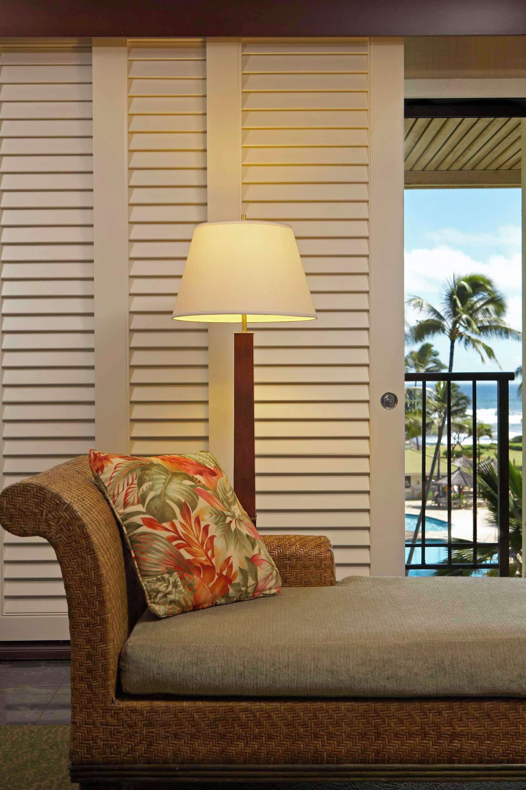 A room with a bed, a lamp, and a view of palm trees and a pool.